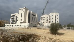 Modern appartementengebouw onder bouw met een skyline van donkere wolken, omgeven door een kale, stoffige grond en enkele jonge bomen, gelegen in een ontwikkelingsgebied nabij Casa la Pluma.