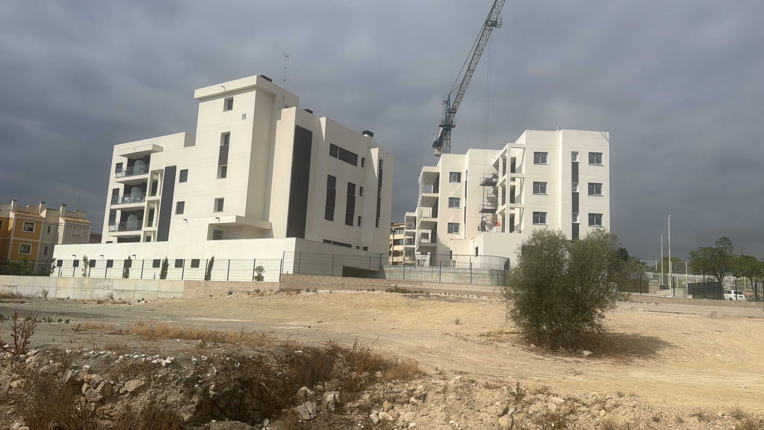 Modern appartementengebouw onder bouw met een skyline van donkere wolken, omgeven door een kale, stoffige grond en enkele jonge bomen, gelegen in een ontwikkelingsgebied nabij Casa la Pluma.