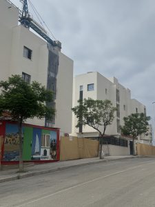 Modern woningbouw in aanbouw met strakke, witte gevels en grote ramen, omgeven door bomen en een bouwhek met promotionele afbeelding, gelegen in een stedelijke omgeving.